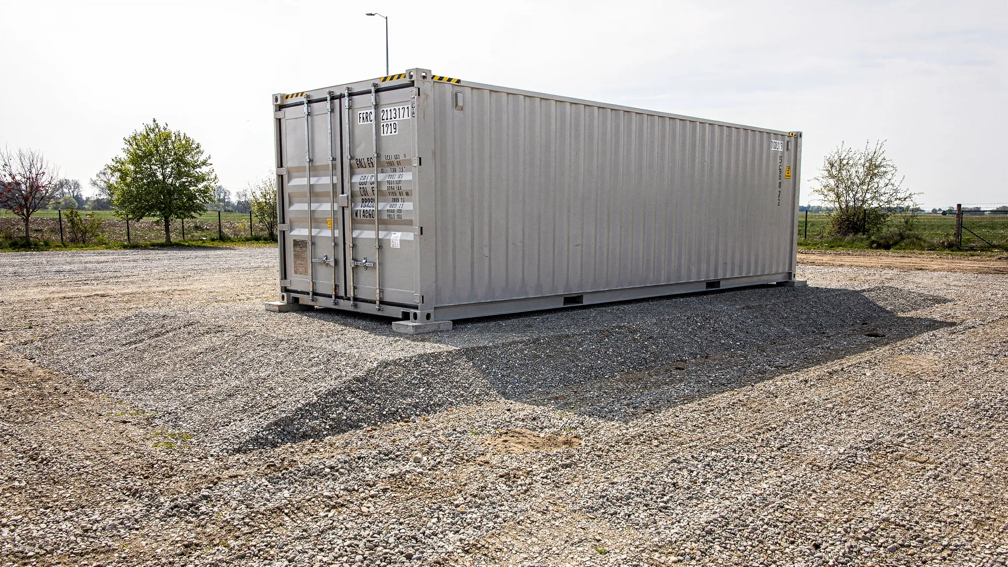 A shipping container placed on a compacted gravel pad with corner blocks, showing level placement, drainage slope away from the container, and clear access space for a delivery truck.