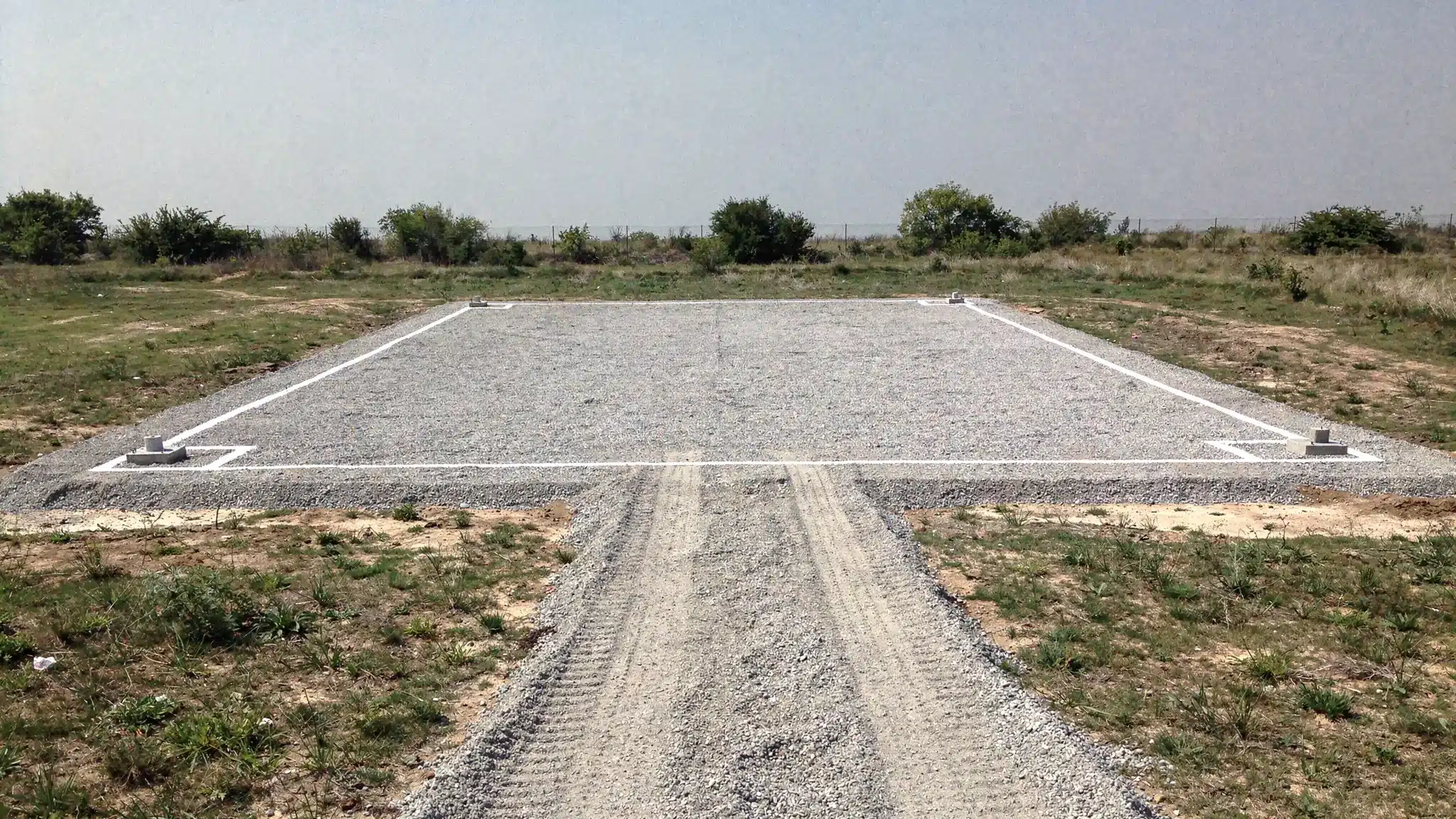 Container Solutions for Jobsites, Farms, and Small Business A simple site preparation scene showing a compacted gravel pad with four corner supports marked for a 40ft container, with a clear truck approach path and no overhead obstructions.