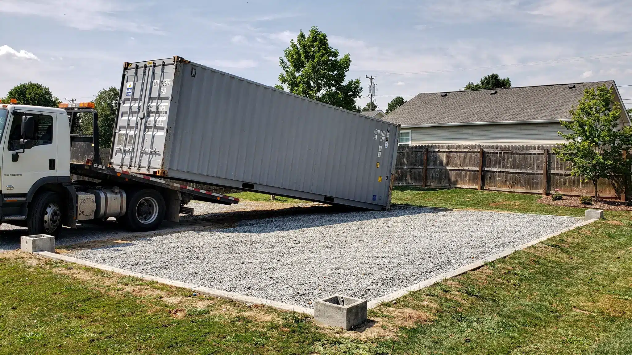 A steel ISO shipping container being delivered on a tilt-bed truck onto a level gravel pad at a suburban Raleigh, North Carolina property. The truck is angled for offload, with clear overhead clearance and visible corner blocks marking the placement spot.