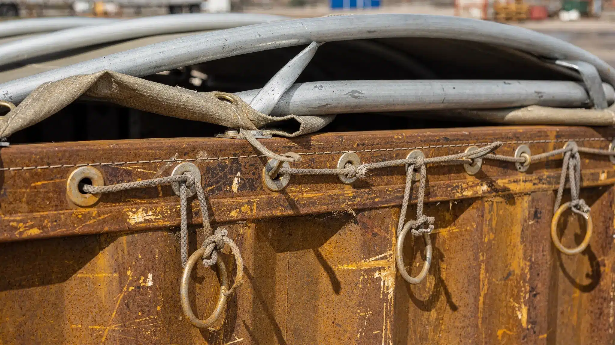 Open Top Container Guide: Loading, Tarps, and Common Uses Close-up of an open top container’s tarp system showing roof bows, tarp edge with grommets, and rope or strap tie-downs along the top rail, with the texture of Corten steel visible.