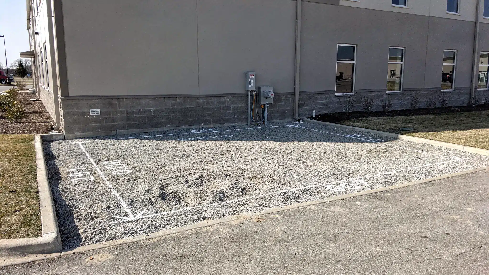 A prepared gravel pad beside a commercial building with a clearly marked container footprint, a nearby outdoor electrical disconnect, and a clear access path for a tilt-bed delivery truck.