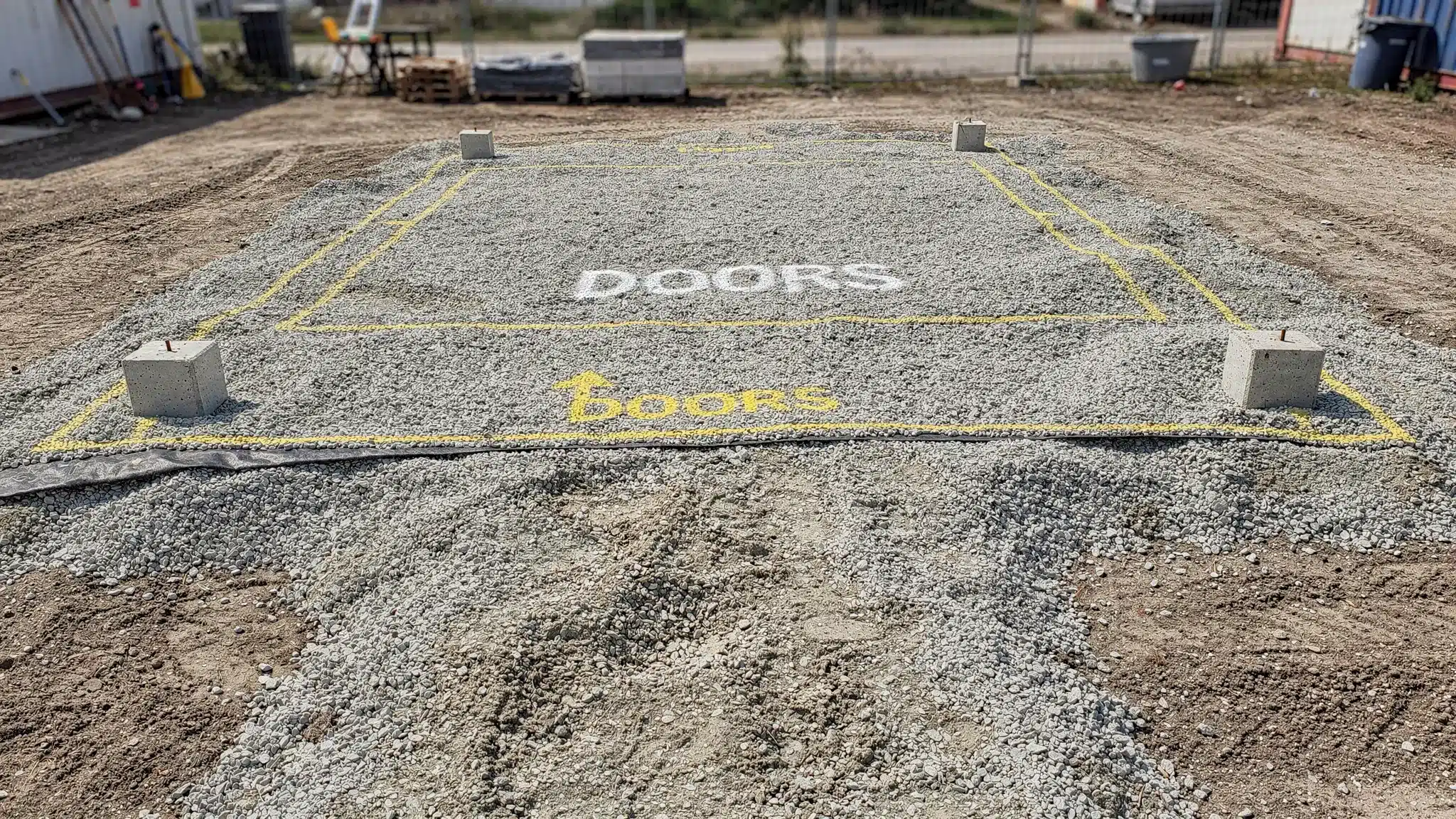 A prepared shipping container pad made of compacted gravel with geotextile underlayment, four corner support blocks, clear access path, and spray-painted footprint markings showing door orientation.