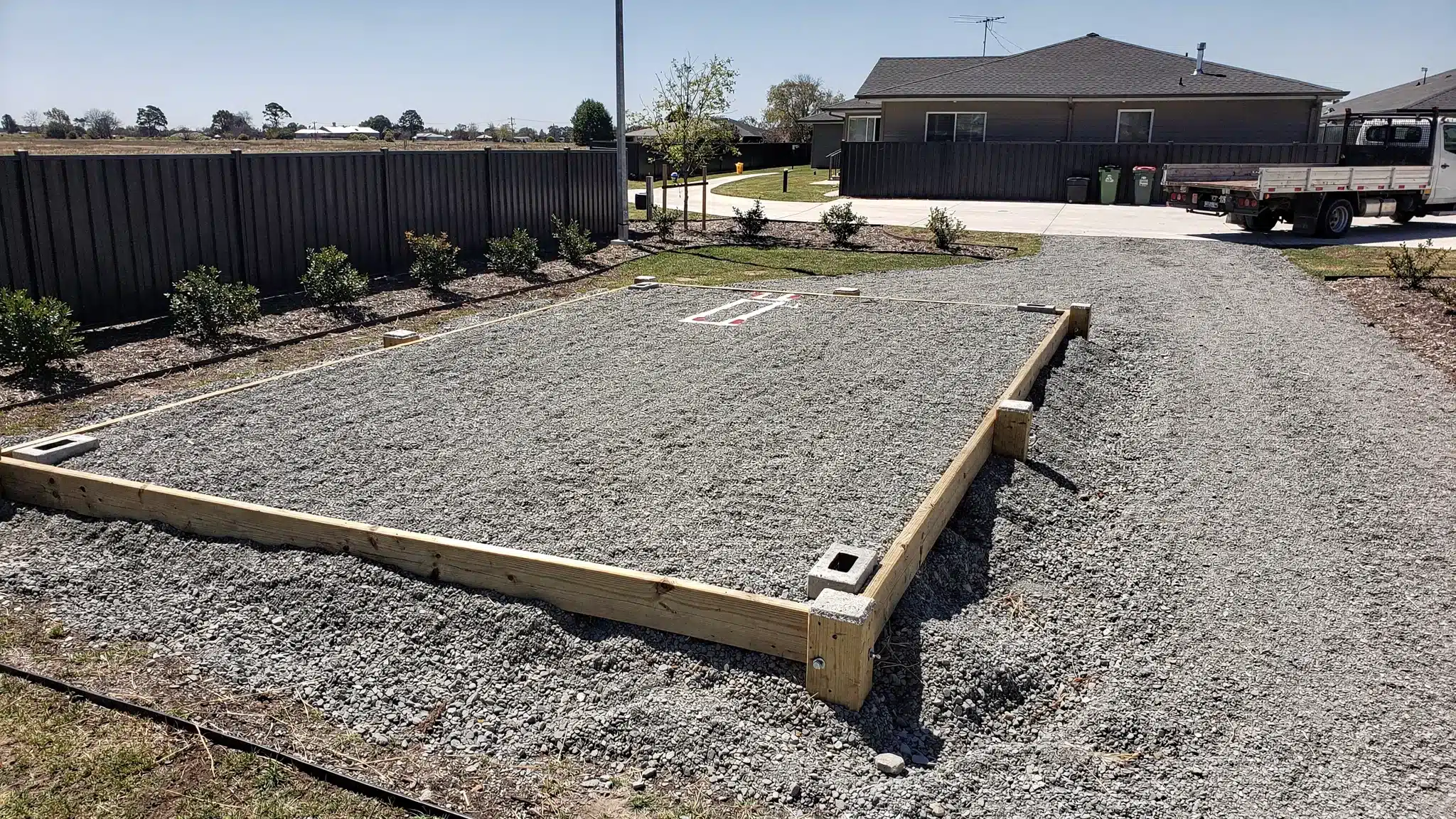 A properly prepared container pad with compacted gravel, level corner supports, and clear access path for a tilt-bed truck. The scene shows a residential or light commercial property with marked drop zone, good drainage slope, and no overhead obstructions.