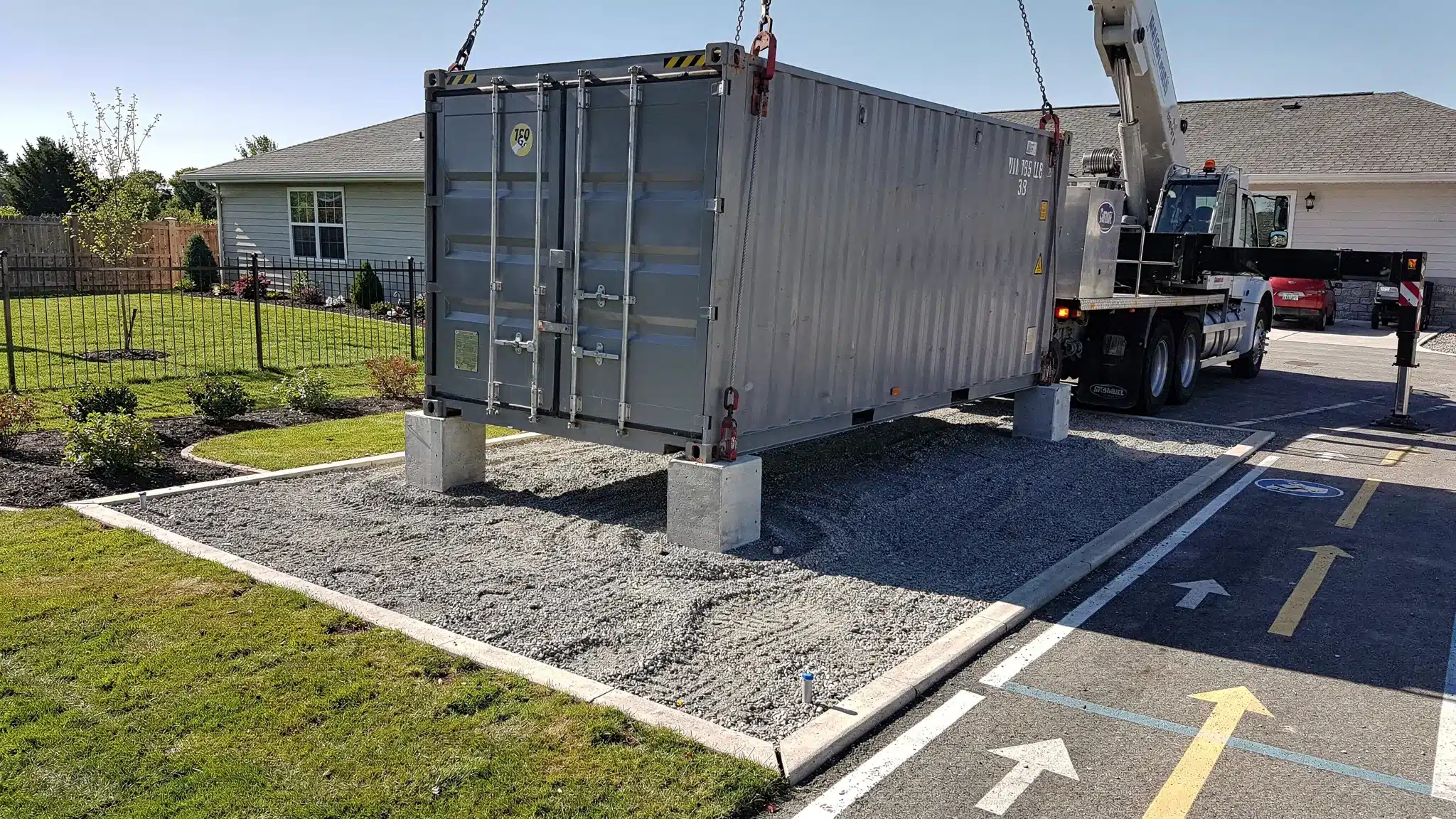 A realistic outdoor scene showing a shipping container being placed on a compacted gravel pad with corner supports, clear access route markings, and adequate overhead clearance on a residential-style property.