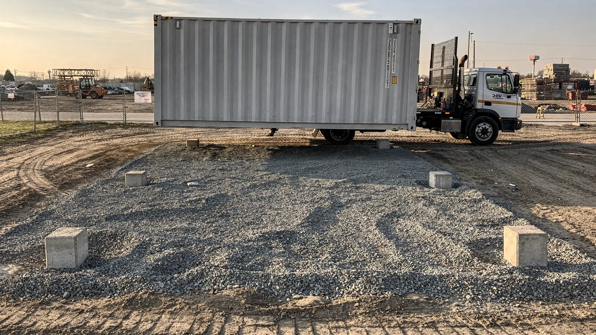 A shipping container being placed on a compacted gravel pad with corner supports at a jobsite, showing a clear delivery path and overhead clearance suitable for a tilt-bed drop.