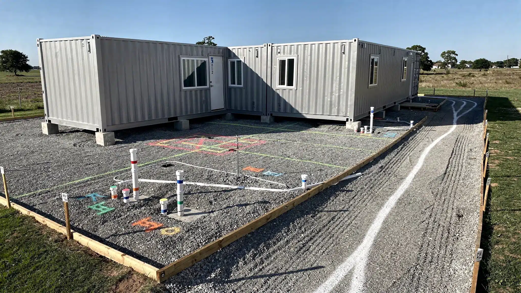 A shipping container home project in progress on a prepared gravel pad, with visible corner blocks/piers, marked utility stub-out locations (water, sewer, electrical conduit), and a delivery path showing turning clearance.