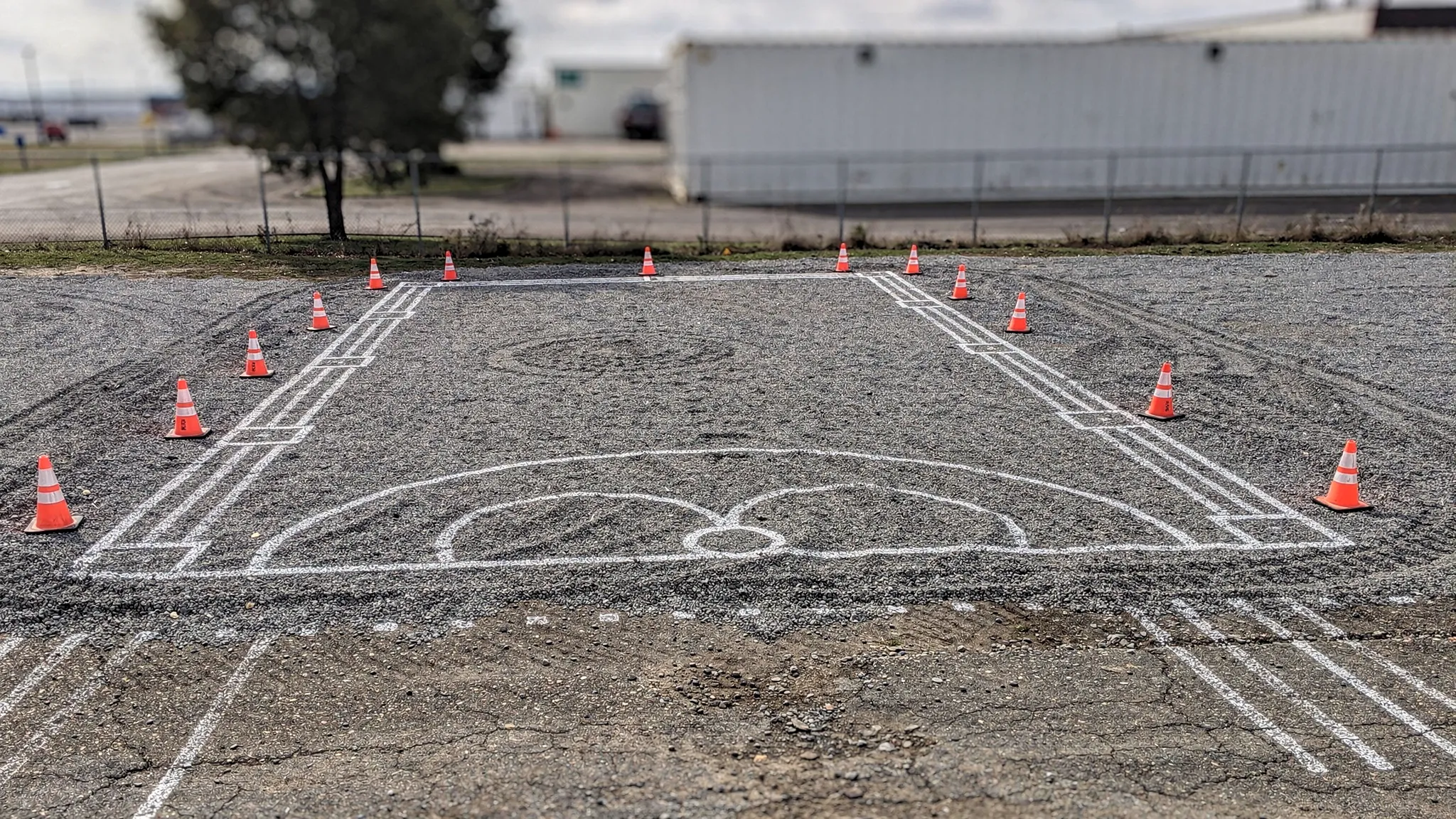 A simple site-planning scene showing a rectangular shipping container footprint on a gravel pad with labeled clearances: door swing zone in front, 3-foot walk path along one side, and a marked truck approach path with turning area. No text overlay, just clear visual cues like cones and chalk lines.