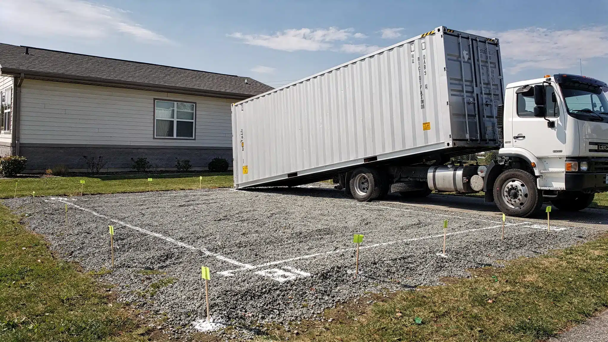 A tilt-bed truck placing a 20ft shipping container onto a compacted gravel pad at a residential or small business property, with clear overhead space and marked corner locations.