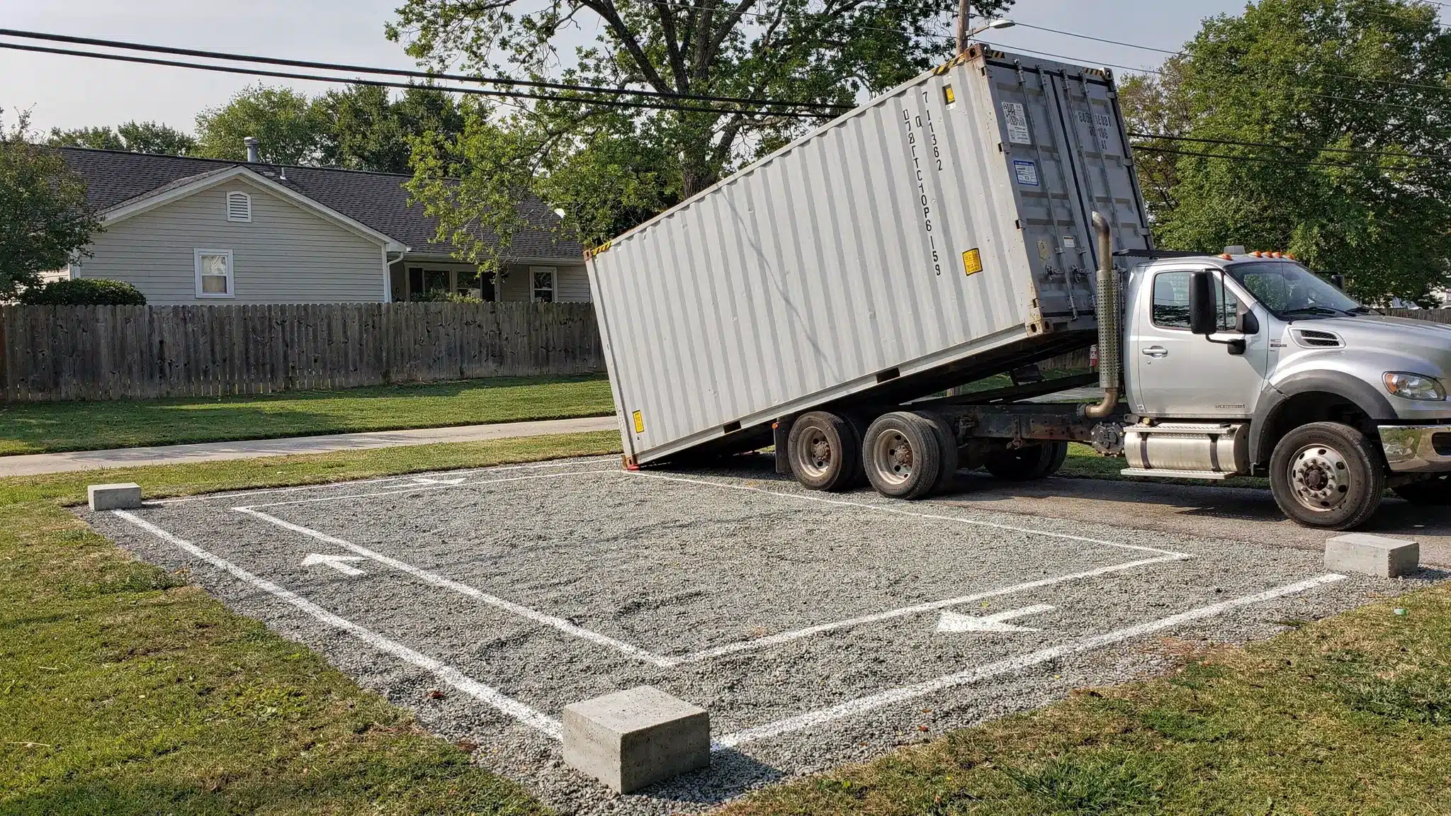 A tilt-bed truck unloading a 20ft shipping container onto a compacted gravel pad on a residential property, with clearly visible corner blocks, a marked drop zone, and plenty of overhead clearance from trees and power lines.