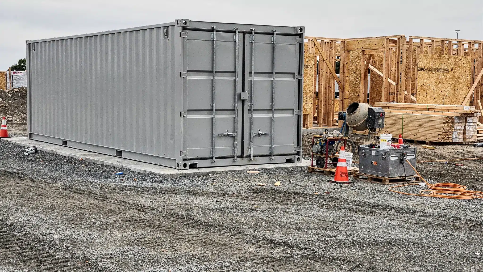 A clean 20 ft shipping container placed on a compacted gravel pad beside a small construction site, with tools and materials organized nearby and clear truck access in front of the doors.