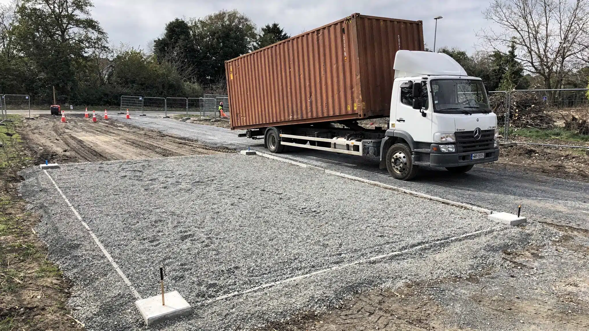 What to Expect From a Storage Container Delivery Service A tilt-bed delivery truck carefully placing a Corten steel shipping container on a level gravel pad at a commercial jobsite, with marked corner locations, clear access, and trees trimmed away from the delivery route.