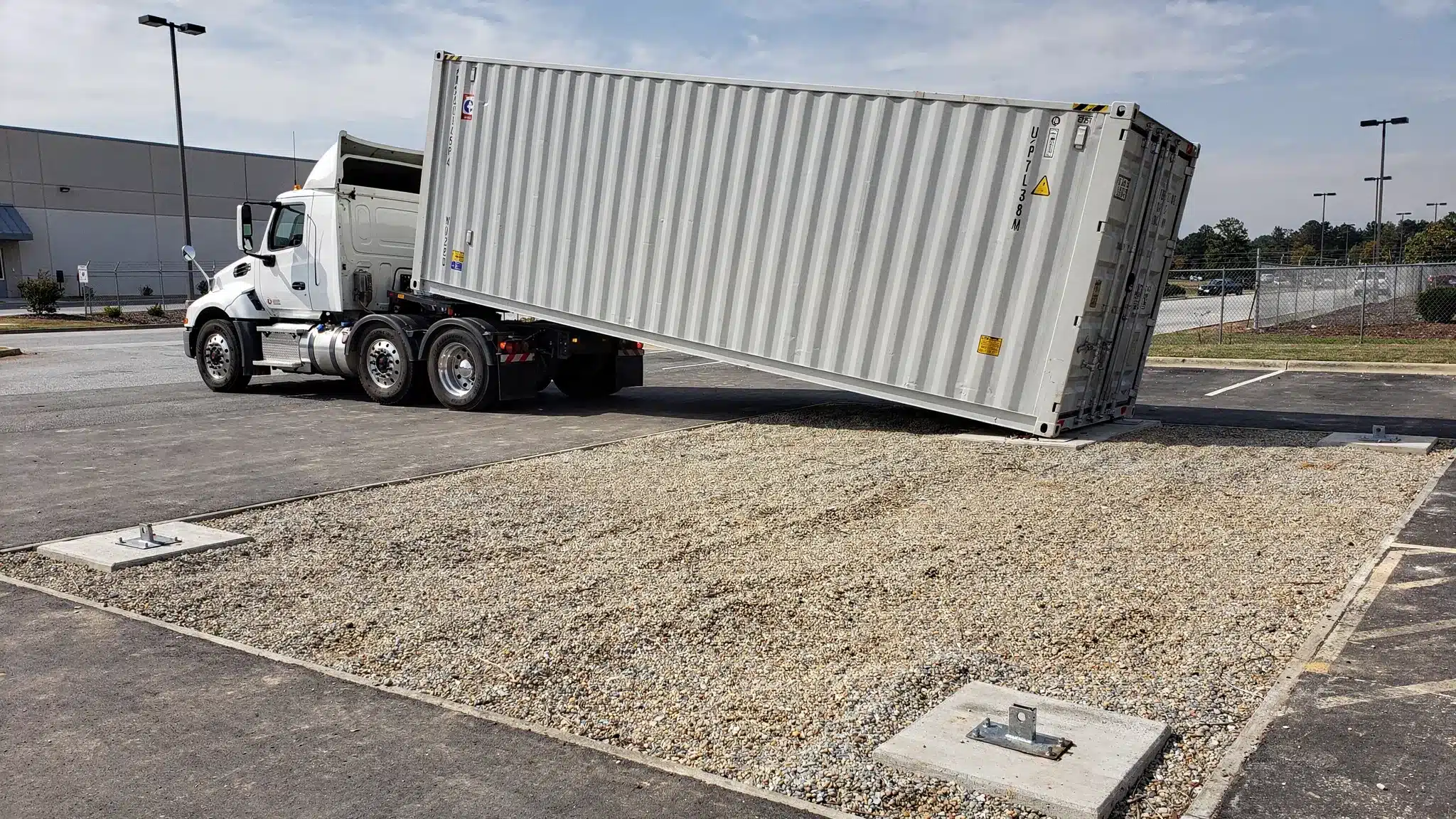 Shipping Container Delivery Cost: What Changes Your Quote Side view of a tilt-bed truck sliding a 40ft High Cube shipping container onto a compacted gravel pad at a commercial property in Raleigh, showing clear driveway access and marked corner support points.