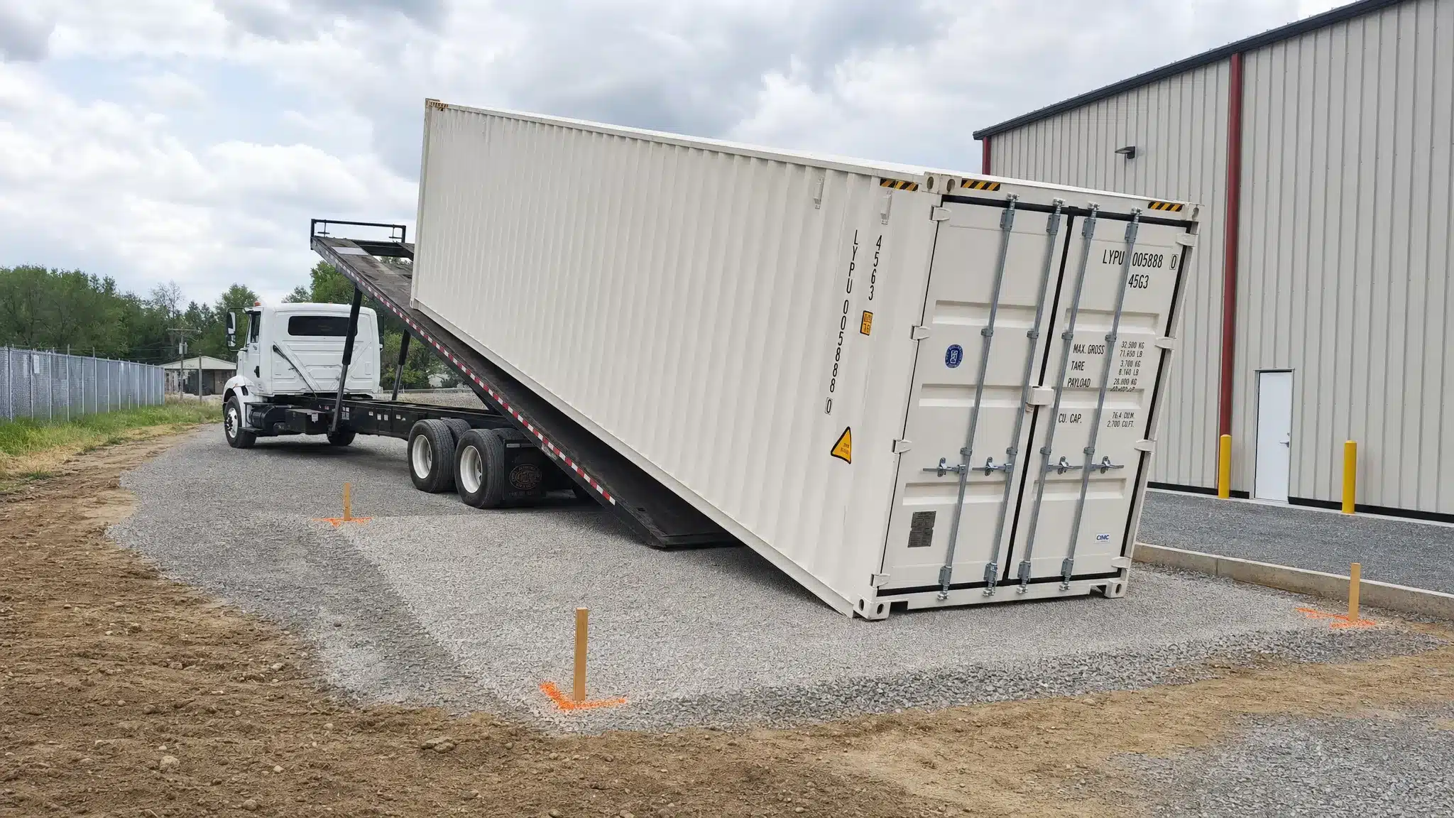 A clean shipping container being set onto a compacted gravel pad at a commercial property, with a tilt-bed truck aligned for drop-off, clear truck access, level ground, and marked corner positions for final placement.