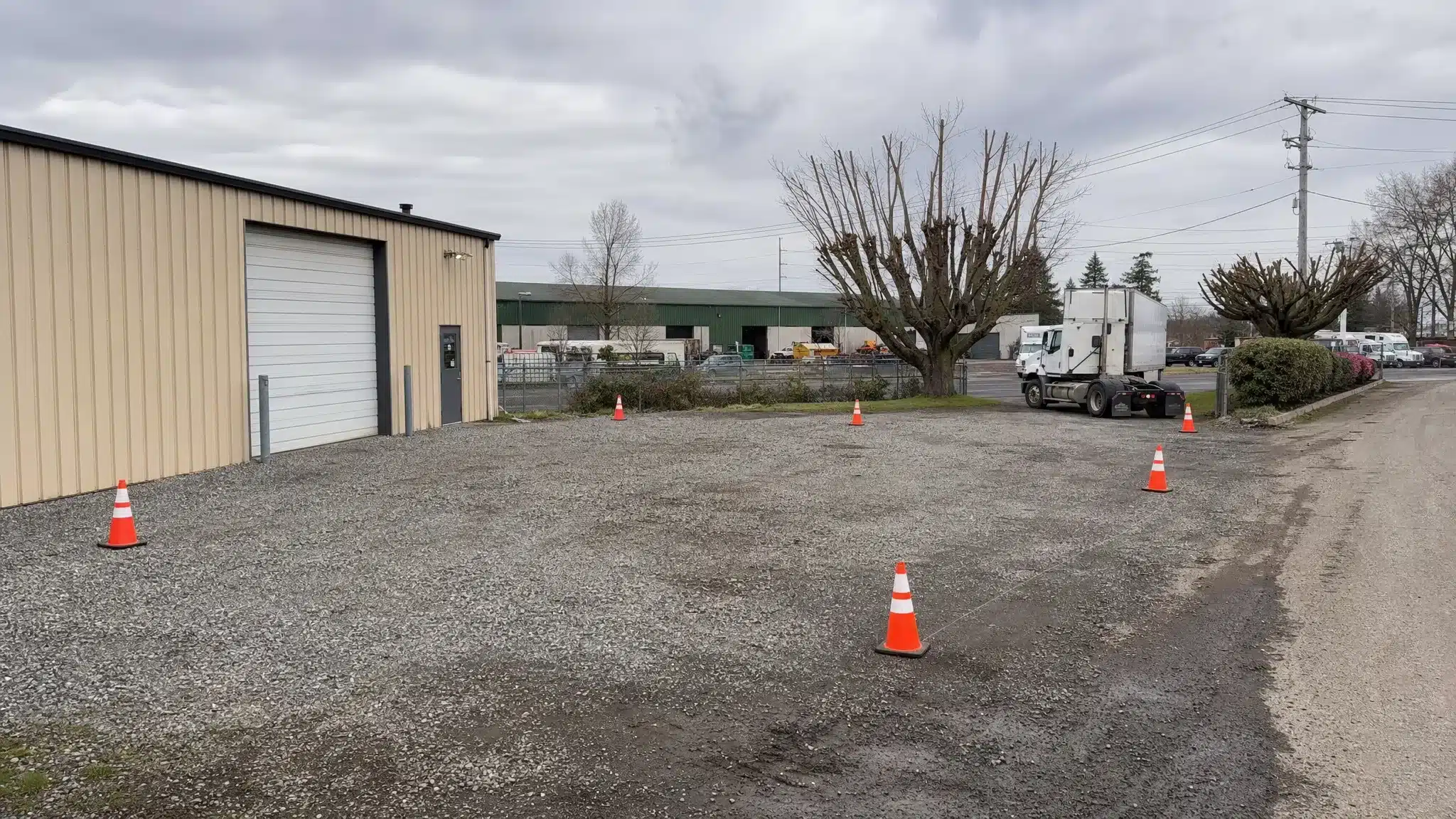 A level gravel pad on a commercial property with cones marking the container footprint, clear truck access, trimmed tree branches, and a ready drop area for a shipping container delivery.