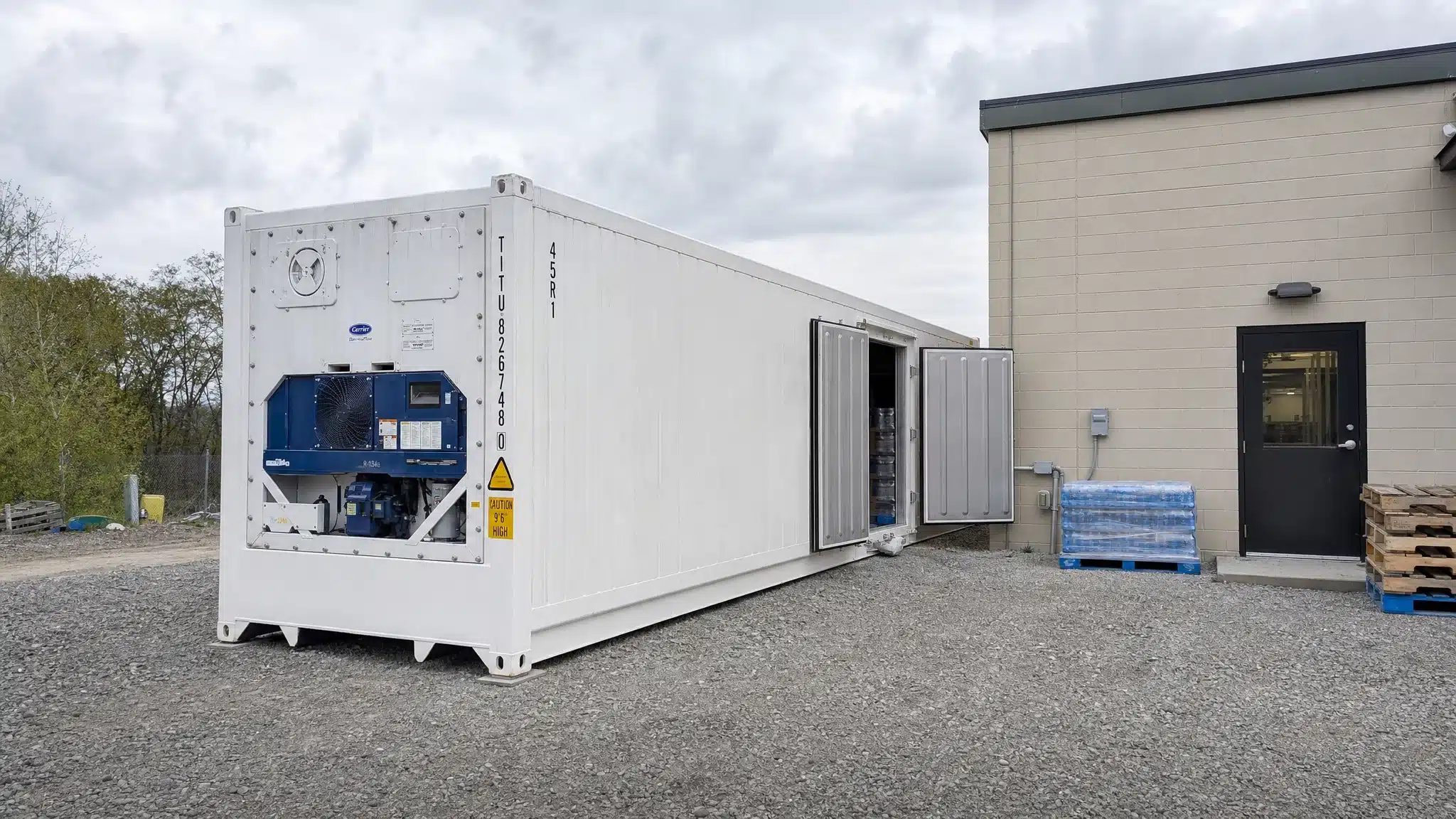 A refrigerated shipping container beside a small commercial building, with clear service space around the refrigeration unit, pallets organized near the open doors, and the unit set on a level gravel pad.