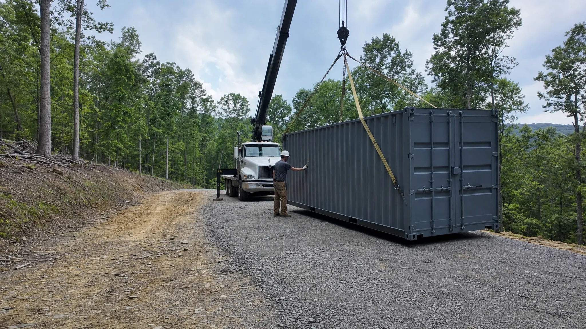 A shipping container being set onto a level gravel pad beside a cleared access path, with open space for the delivery truck and no overhead obstructions.