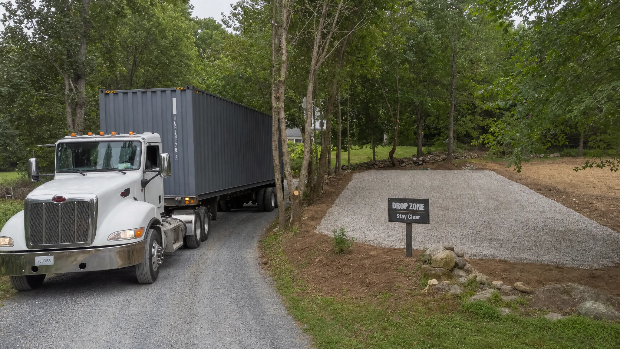 Delivering a Shipping Container to Tight Access Sites A shipping container delivery truck maneuvering beside a narrow gravel driveway with trees trimmed back, a marked drop zone, and a level gravel pad ready for placement.