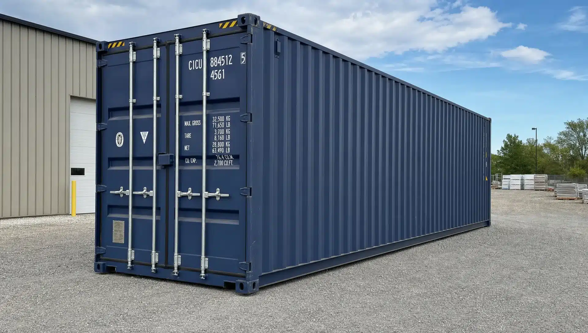 A 40ft standard shipping container resting on a level gravel pad at a commercial property, shown from a low three-quarter front angle with closed cargo doors, visible corner castings, and open space around the delivery lane for truck access.